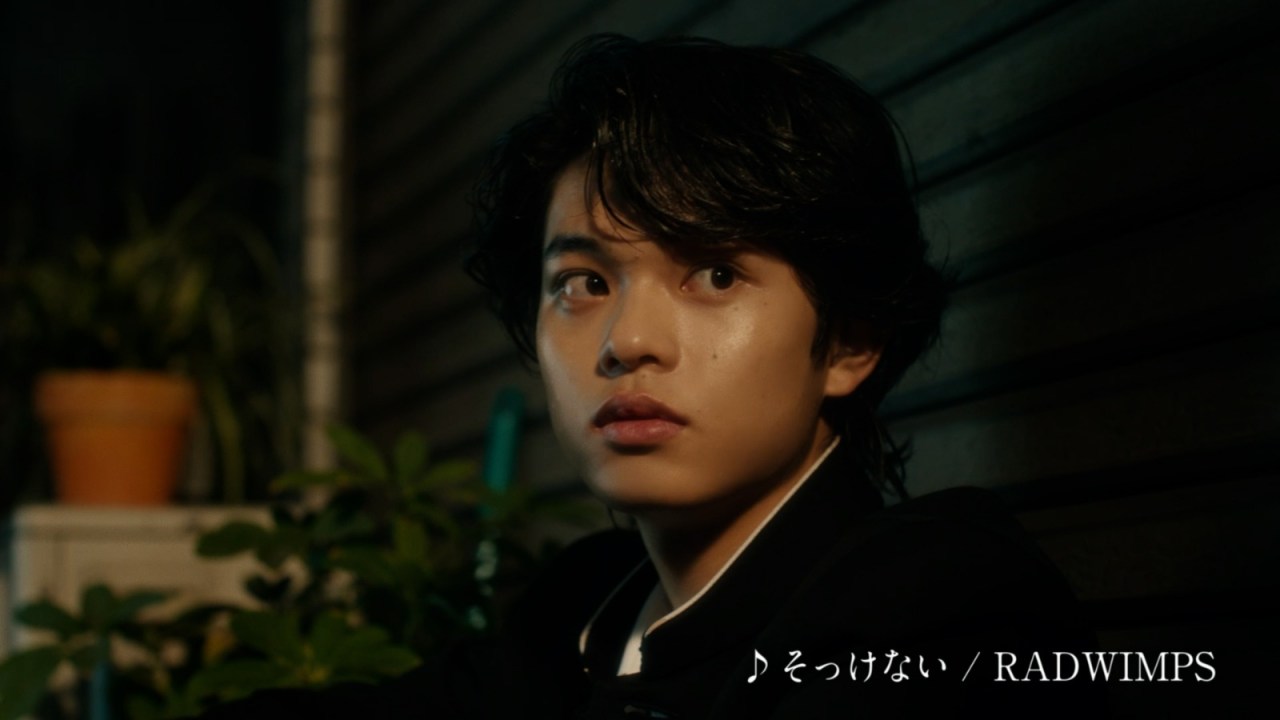 A young person with dark, wavy hair is sitting against a wall, looking to the side with a surprised expression. They are wearing a black outfit and there are potted plants in the background. The text at the bottom indicates a song title by RADWIMPS.