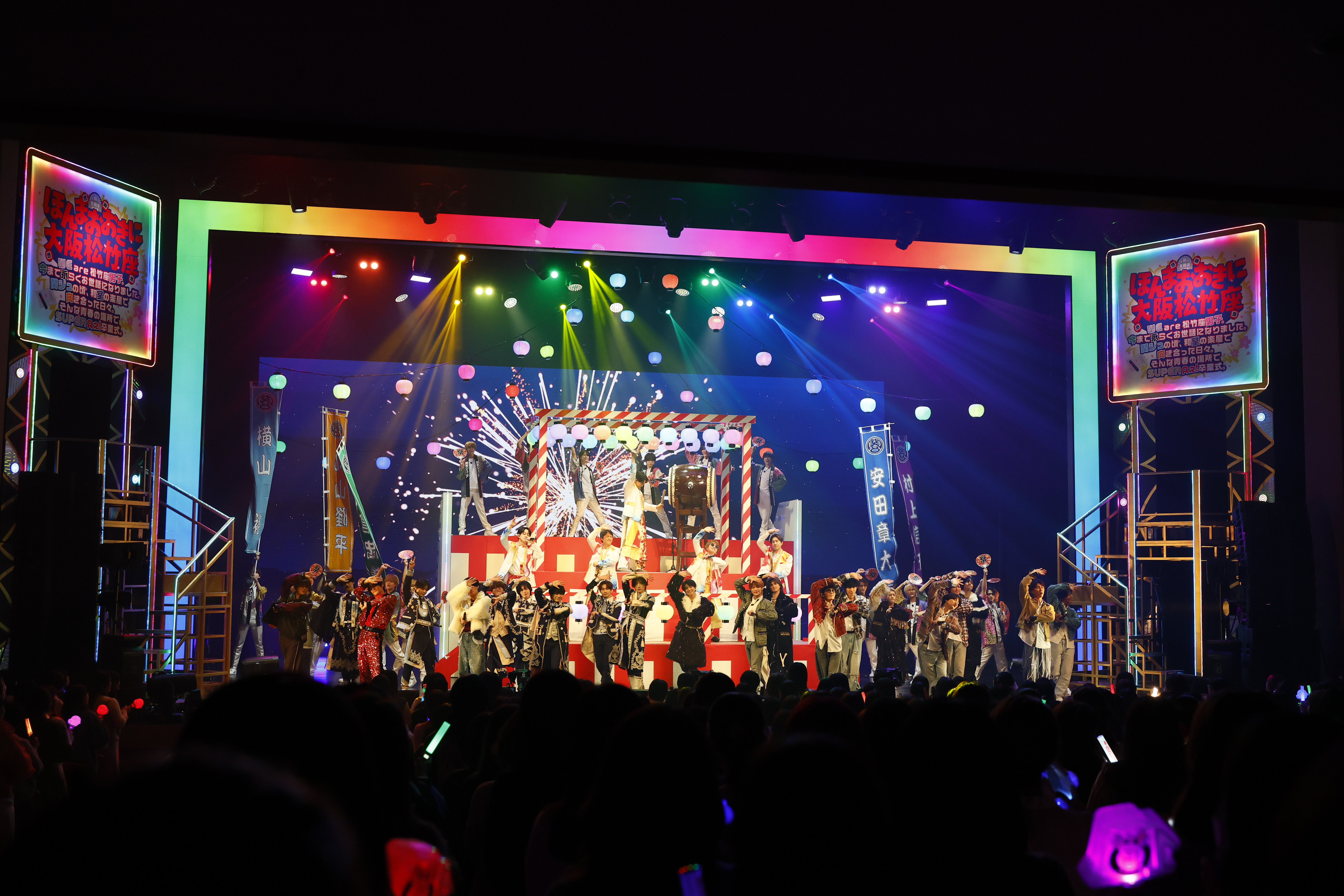 A vibrant stage performance featuring multiple performers in colorful costumes, surrounded by festive lighting and decorations. The background displays a colorful graphic with fireworks, and the audience is visible, holding light sticks.