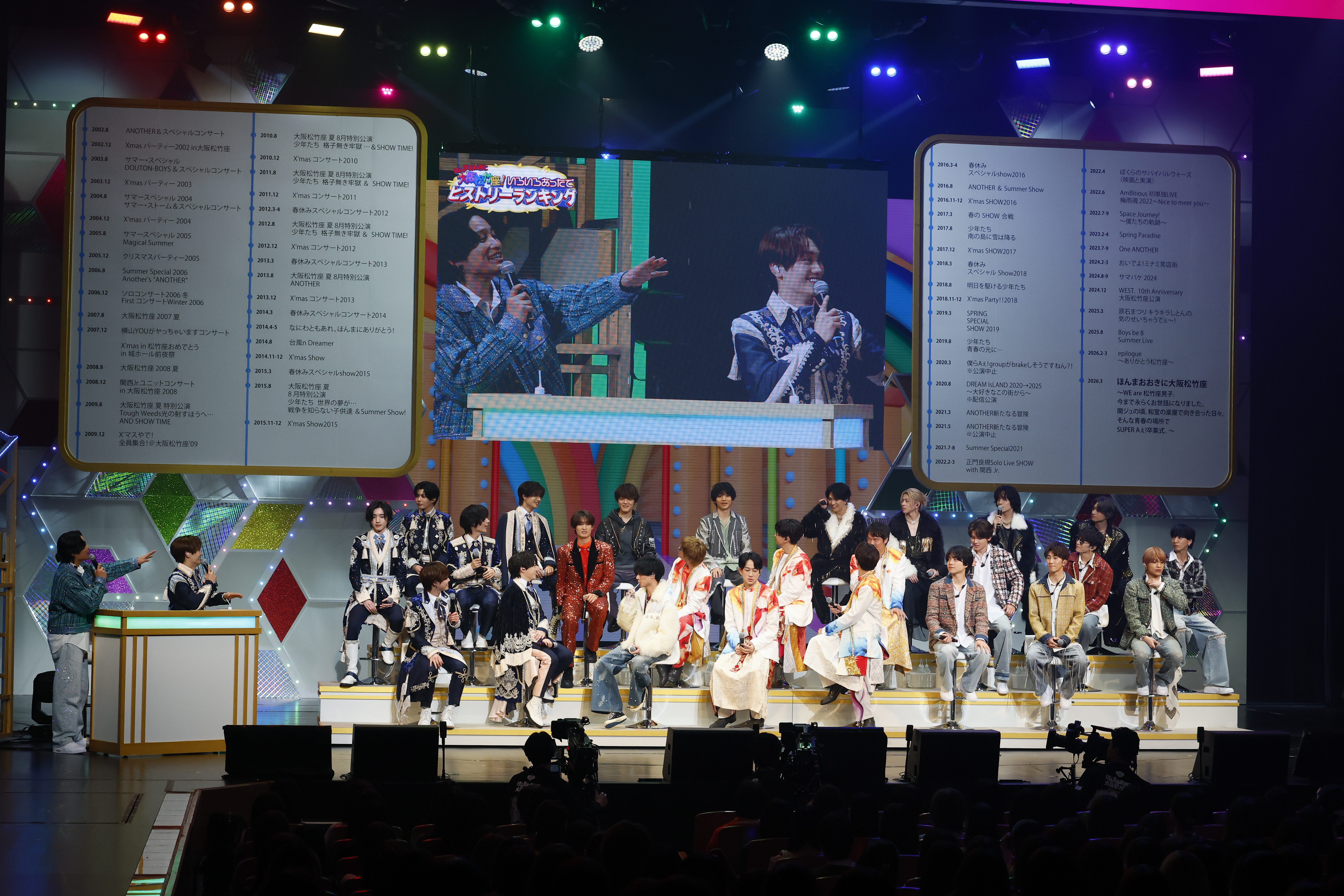 A live performance event featuring a group of performers on stage, with one performer speaking into a microphone while pointing, and a large screen displaying text and images in the background. The audience is visible in the foreground, with colorful lights illuminating the stage.