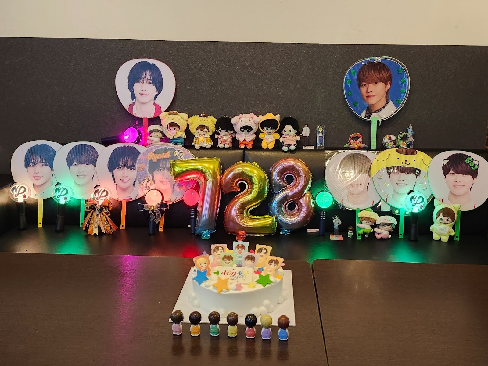 A festive display featuring birthday decorations, including photos of a person, colorful balloons shaped like the numbers 7 and 2, lightsticks, plush toys, and a cake adorned with stars and figures.