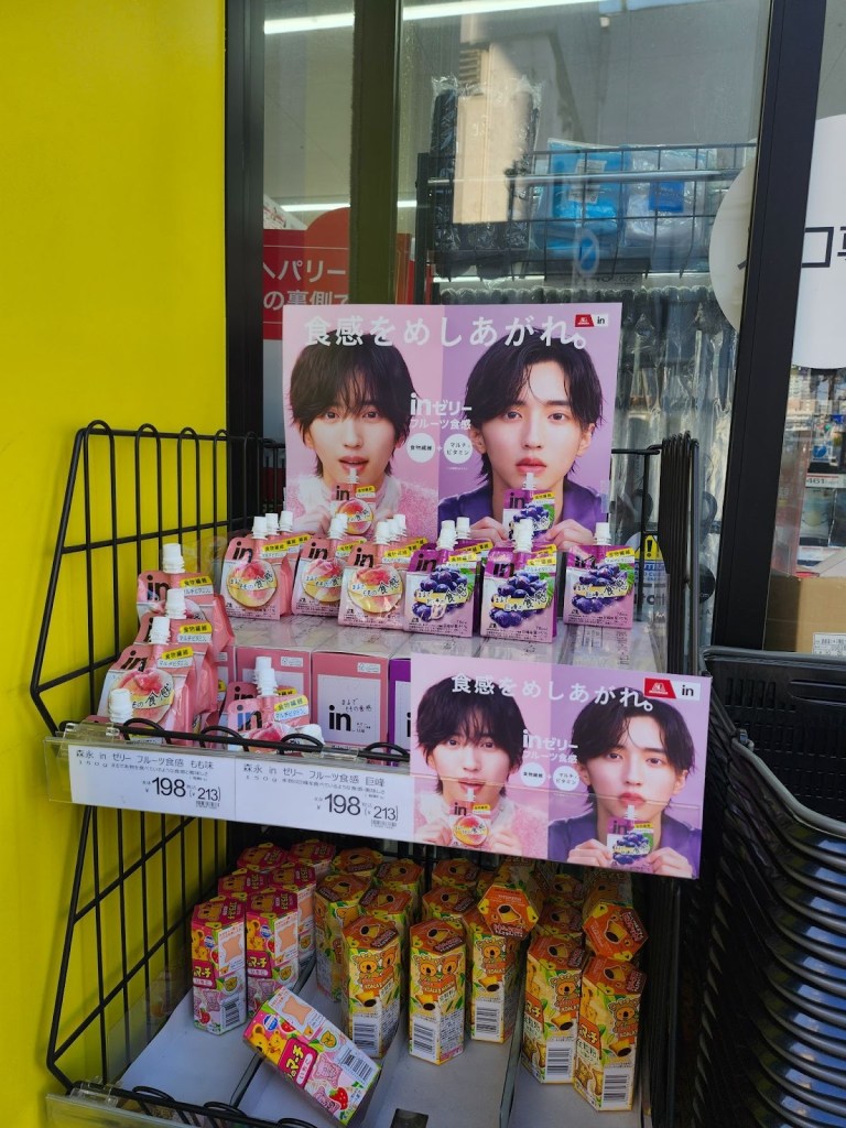 Store display featuring various flavors of jelly drinks with promotional posters of two young men, surrounded by colorful juice boxes and set against a bright yellow background.