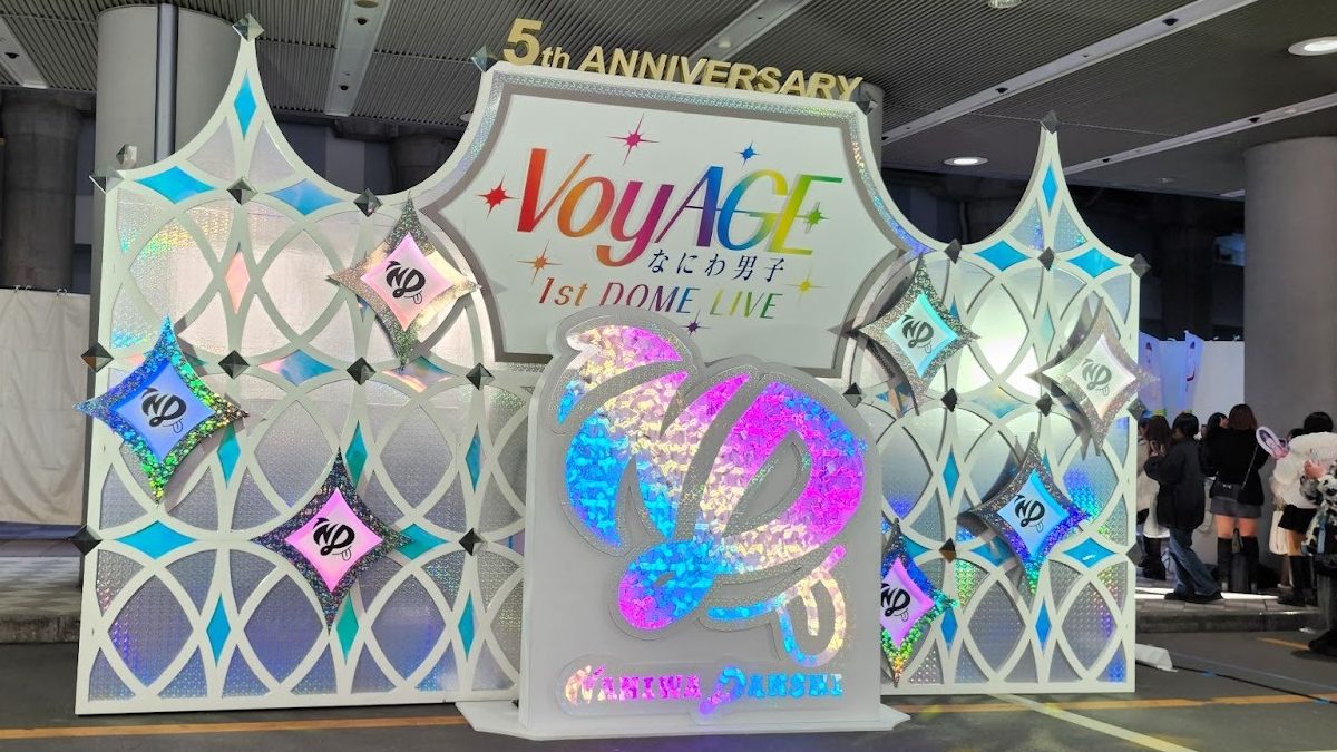 A decorative backdrop featuring the text 'VoyAGE 5th ANNIVERSARY 1st DOME LIVE' in colorful letters, surrounded by shimmering diamond shapes and iridescent designs. The backdrop is designed for a celebration event.