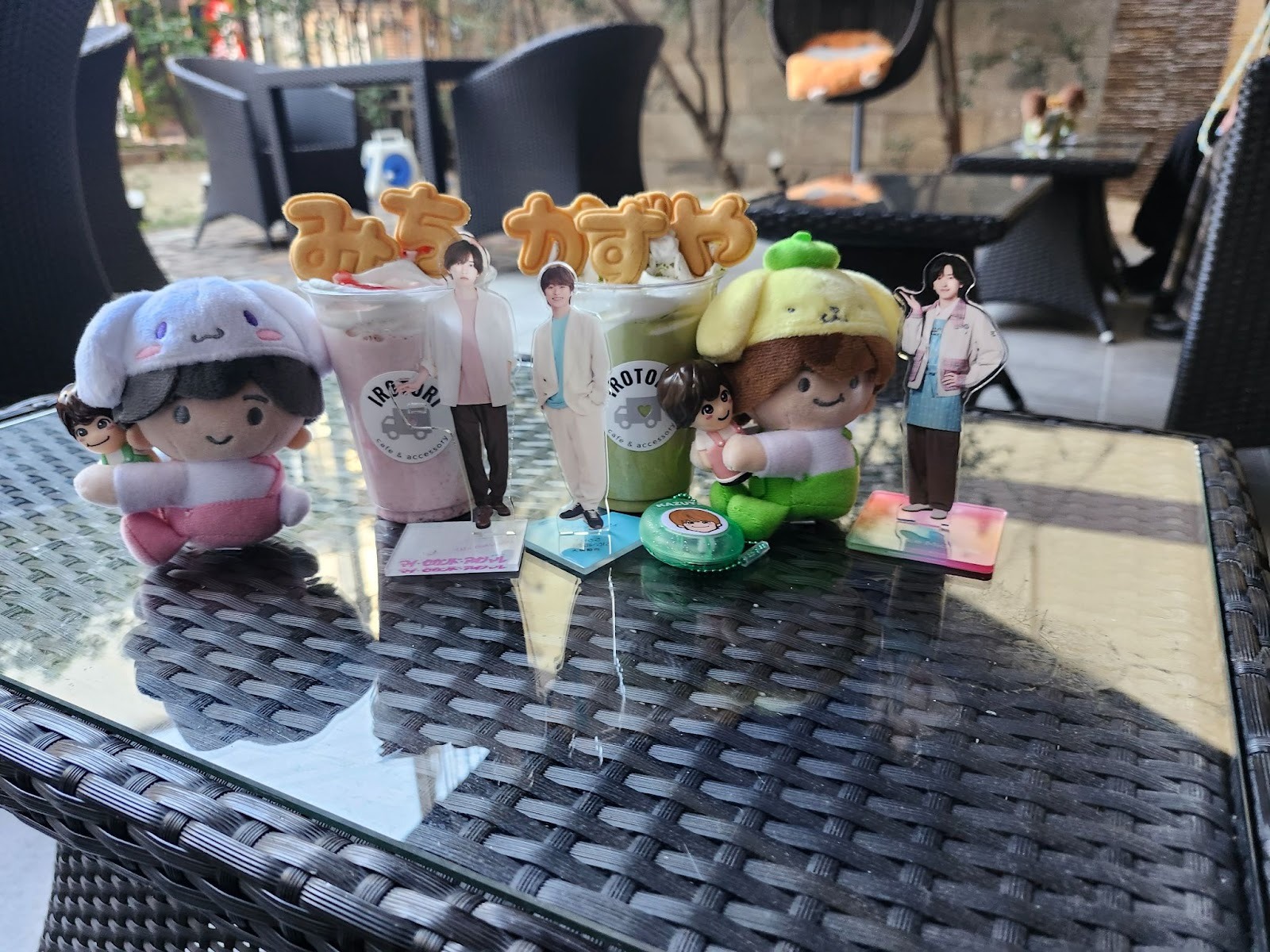 A colorful tabletop display featuring cute plush toys and standees beside drinks topped with decorative cookies. The scene is set outdoors with a textured wicker surface.