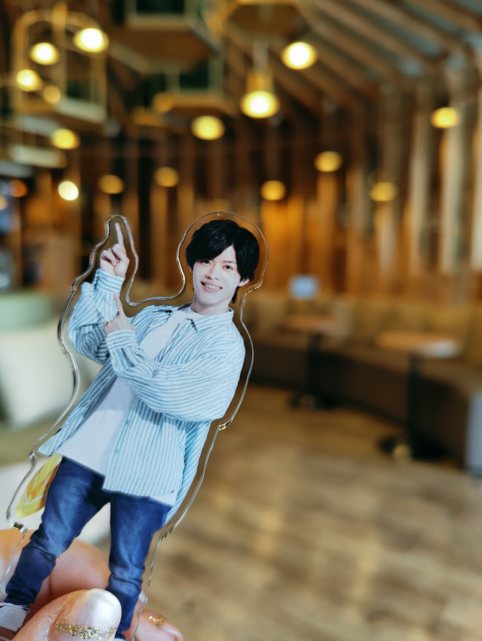 A hand holding a cut-out figure of a young man in a striped shirt, smiling and giving a thumbs up, set against a softly blurred background of a modern cafe interior.