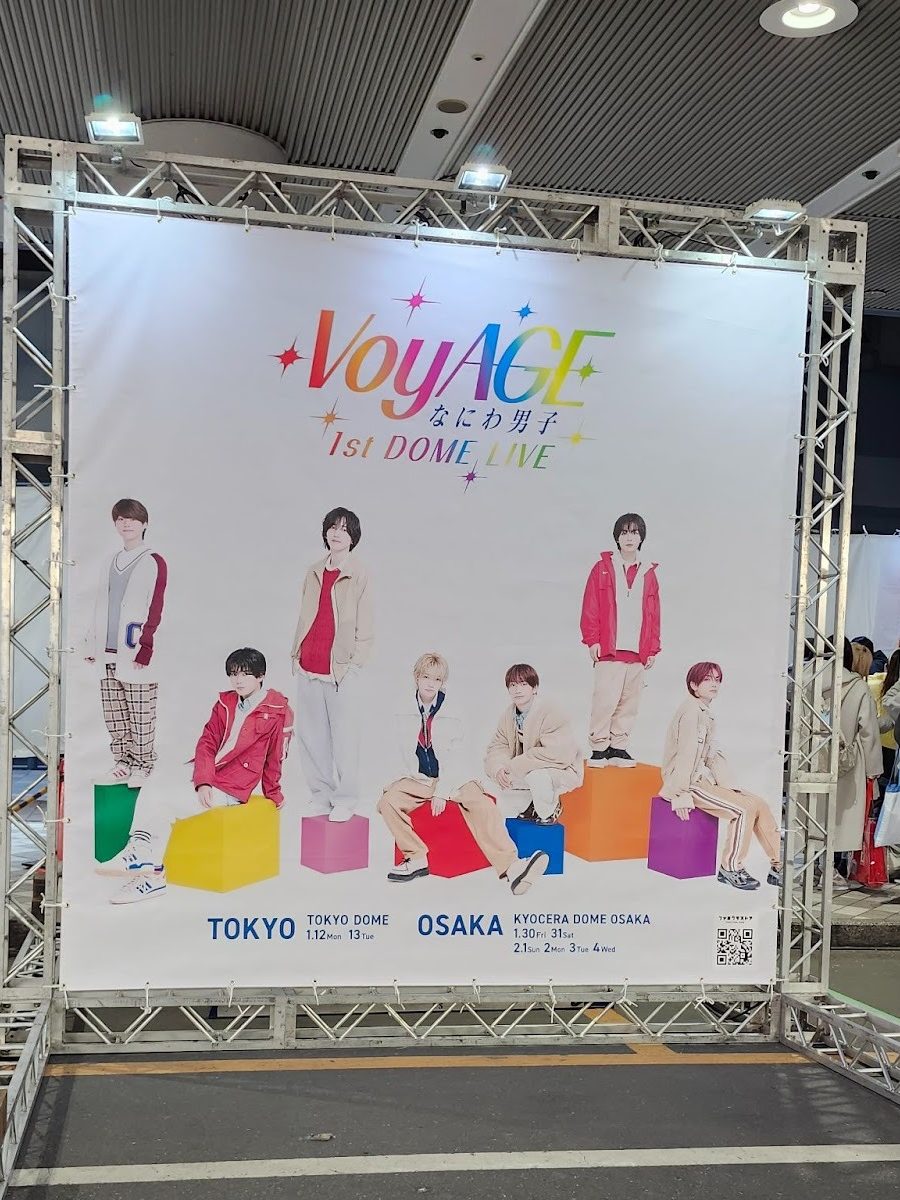 Promotional poster for VoyAGE 1st Dome Live featuring eight members in colorful outfits, standing and sitting on colored blocks, with event details for Tokyo and Osaka.
