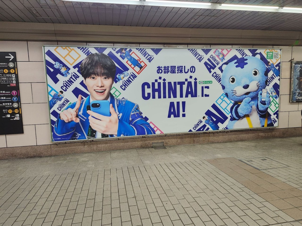 A colorful advertisement featuring a smiling young man holding a smartphone and pointing, alongside a cartoonish blue character, promoting CHINTAI AI. The background includes the text 'CHINTAI!' in bold letters.