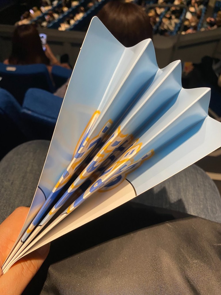 A hand holding a colorful paper fan with blue and yellow designs, in an auditorium setting with blurred audience in the background.
