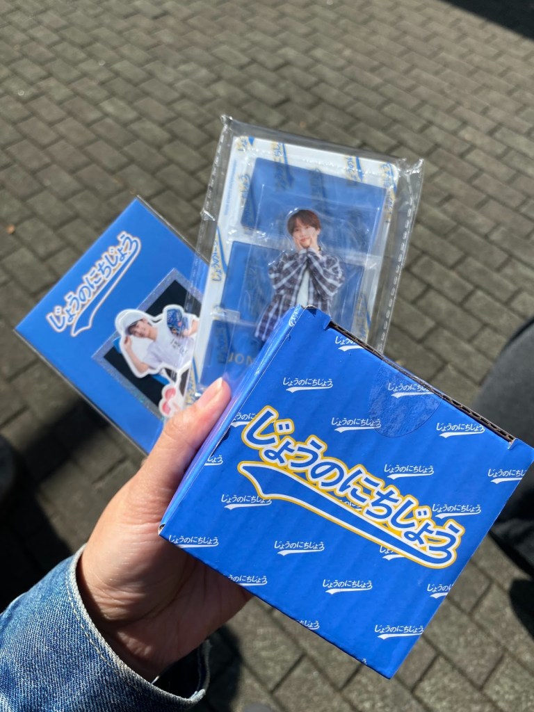 A person's hand holding several items including a blue box and a photocard featuring a young man, with a checkered shirt image visible. The items are branded with Japanese text.
