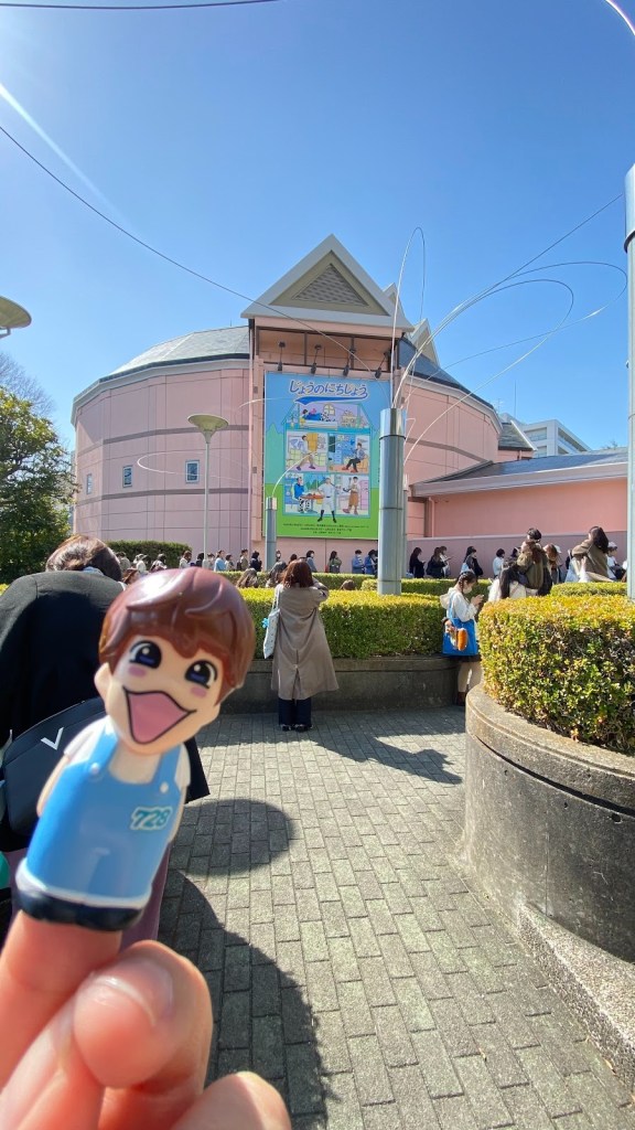 A finger holding a toy figure with a smiling face in front of a pink building featuring a colorful advertisement in a clear blue sky, with a line of people waiting.