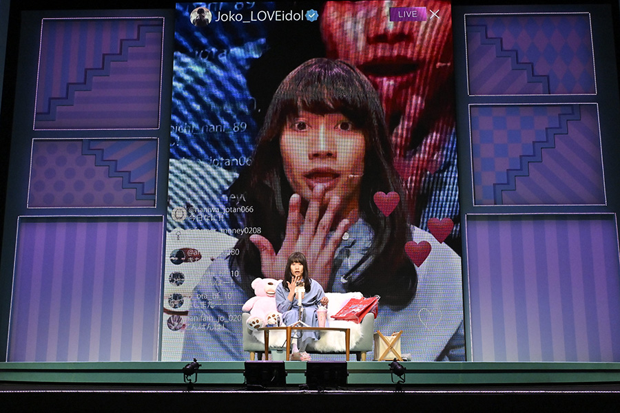A performer sitting on a couch on stage, speaking into a microphone, with a large screen behind displaying their surprised expression and social media interactions, including hearts and user handles.