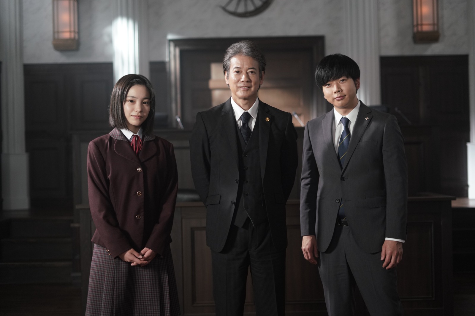 A group of three individuals posing in a courtroom setting, featuring two men in suits and a woman in a school uniform.
