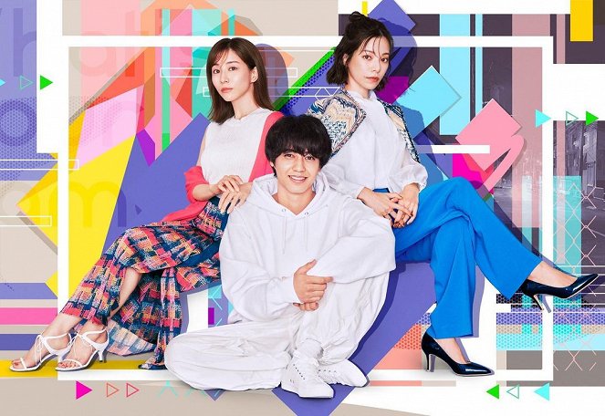 Three young adults posing in a colorful, geometric background. One woman sits on the floor in white attire, flanked by two women standing beside her, one in a patterned skirt and the other in blue pants. They are smiling and facing the camera.