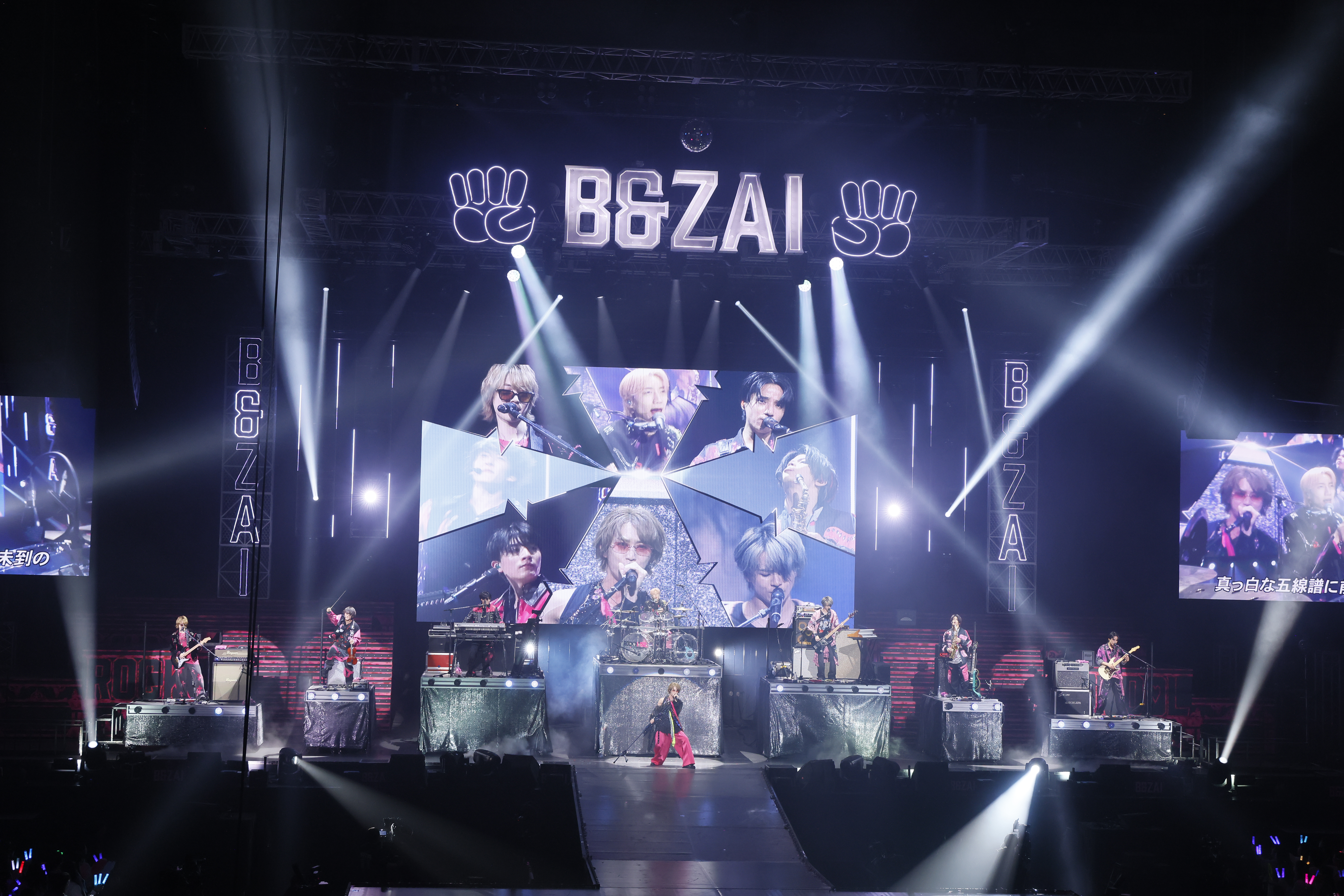 A lively concert scene featuring a band performing on stage with bright lights and large video screens displaying visuals of the artists.