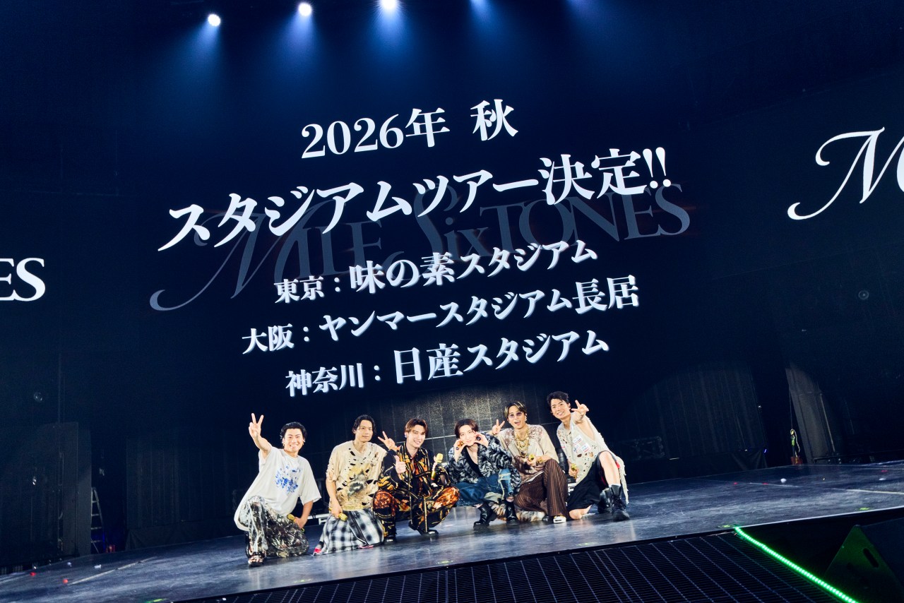 A group of performers posing enthusiastically on stage, with a large screen in the background displaying details about a concert tour scheduled for fall 2026, featuring various venues in Japan.