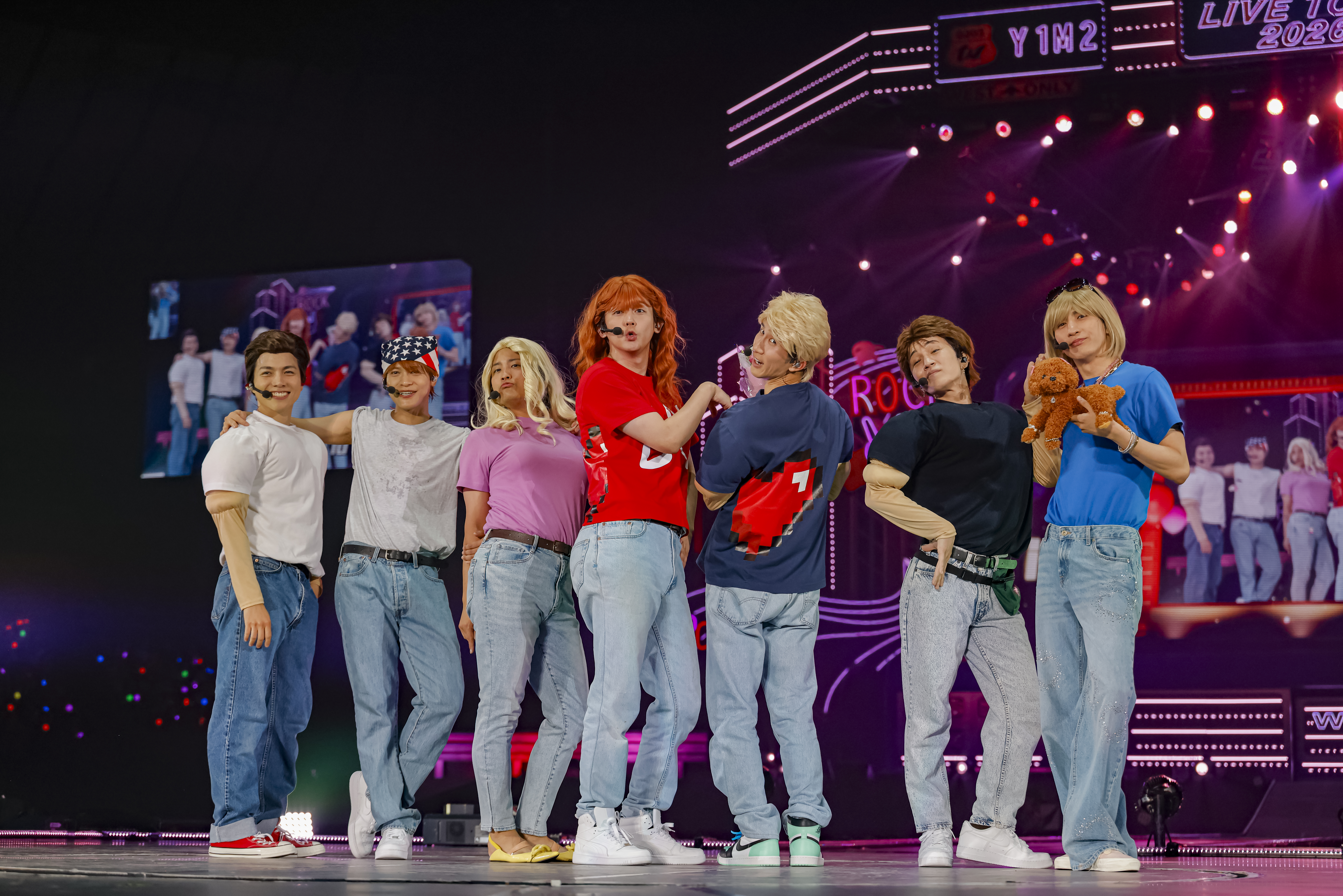 A group of seven performers on stage during a concert, dressed in casual outfits and posing playfully. The backdrop features colorful lights and images of the audience.