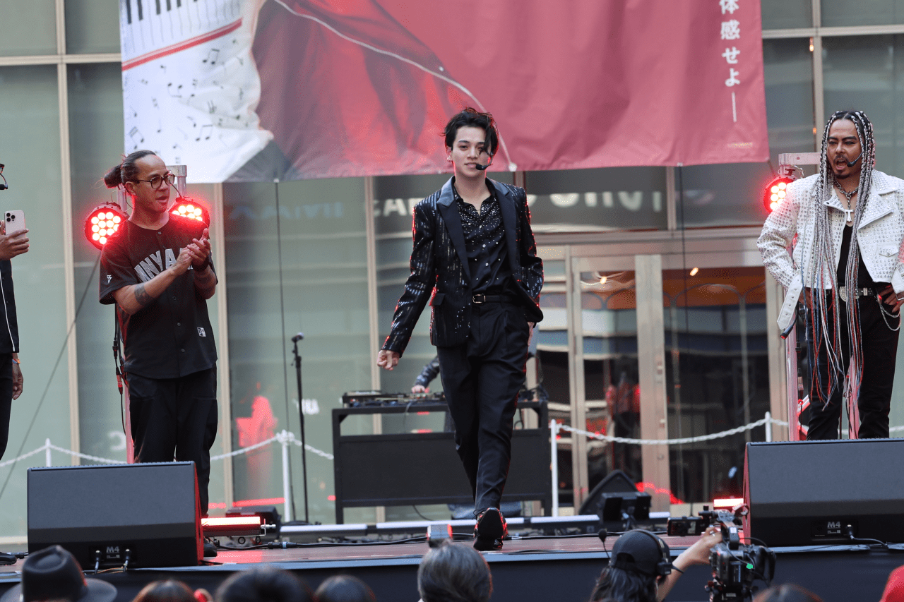 A male performer in a black sequined outfit walks on stage with his microphone, while two other men applaud. The backdrop features musical notes and a vibrant design.