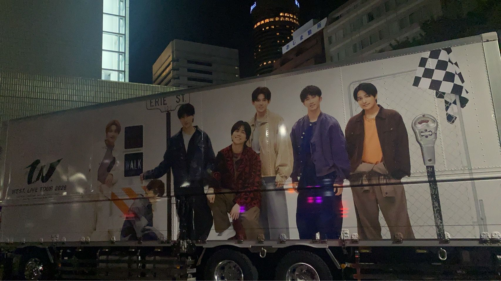 A promotional truck for the West Live Tour 2026 at night featuring five individuals posing in stylish outfits, with a street sign that reads 'ERIE ST.' and a checkered flag draped in the background.