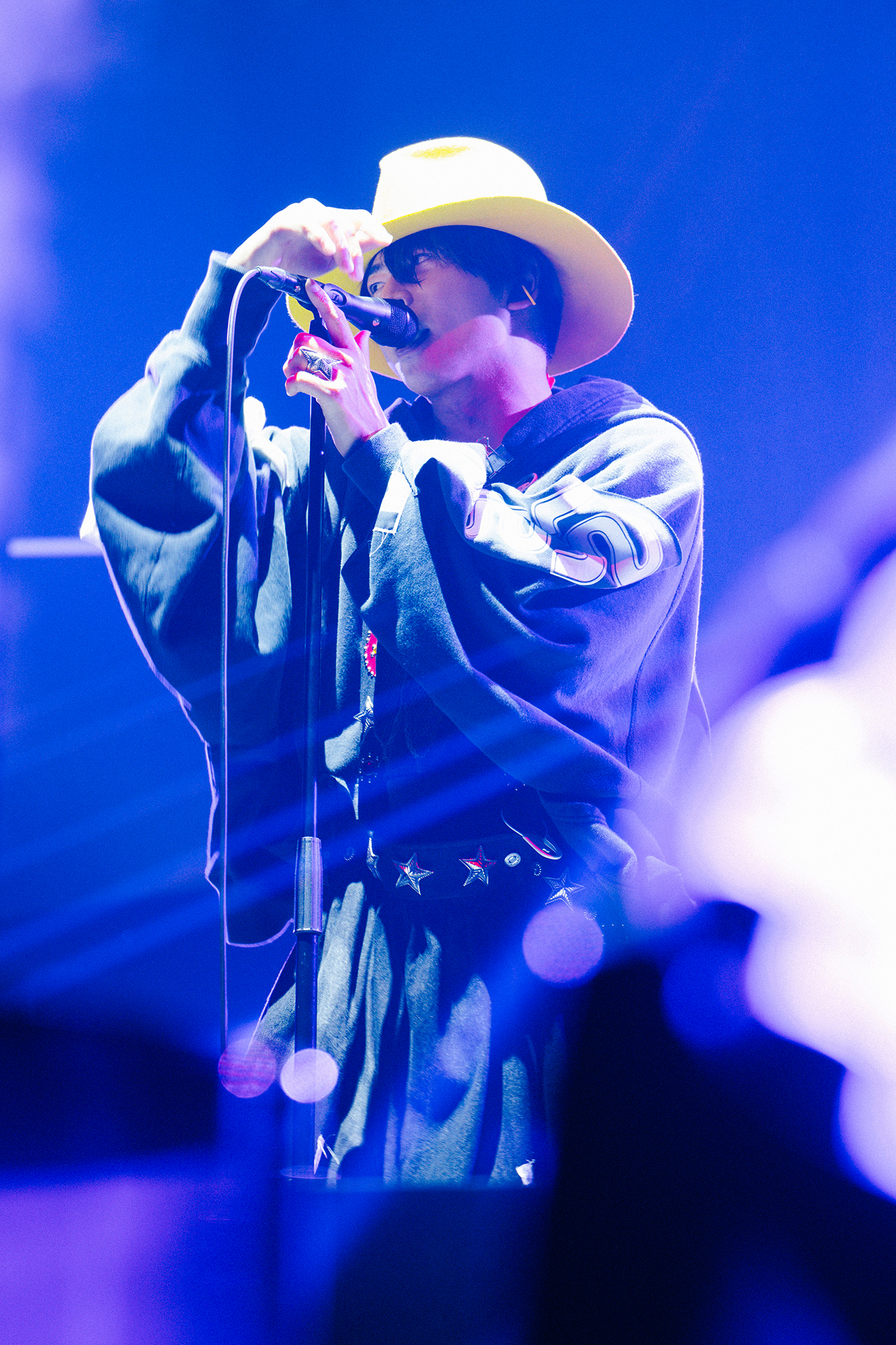 A performer wearing a yellow hat and black hoodie sings into a microphone on stage, with blue lights illuminating the scene.