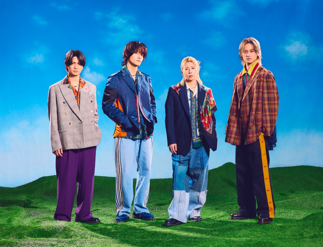Four stylish men stand on a grassy field under a blue sky, wearing unique, colorful outfits that blend modern and vintage elements.
