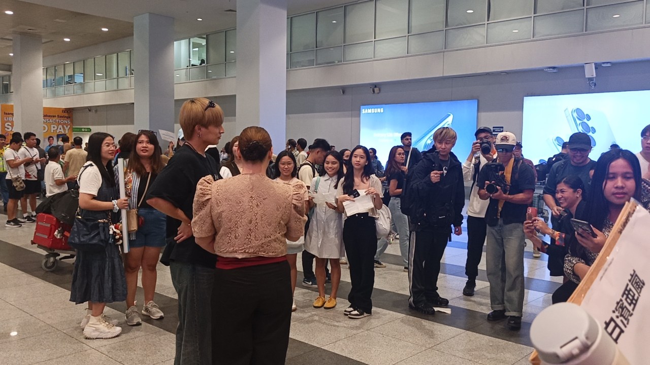 A crowded scene in an airport lobby with a group of fans gathering around two individuals, one dressed in black and another in a light-colored top, while others hold cameras and papers, capturing the moment.