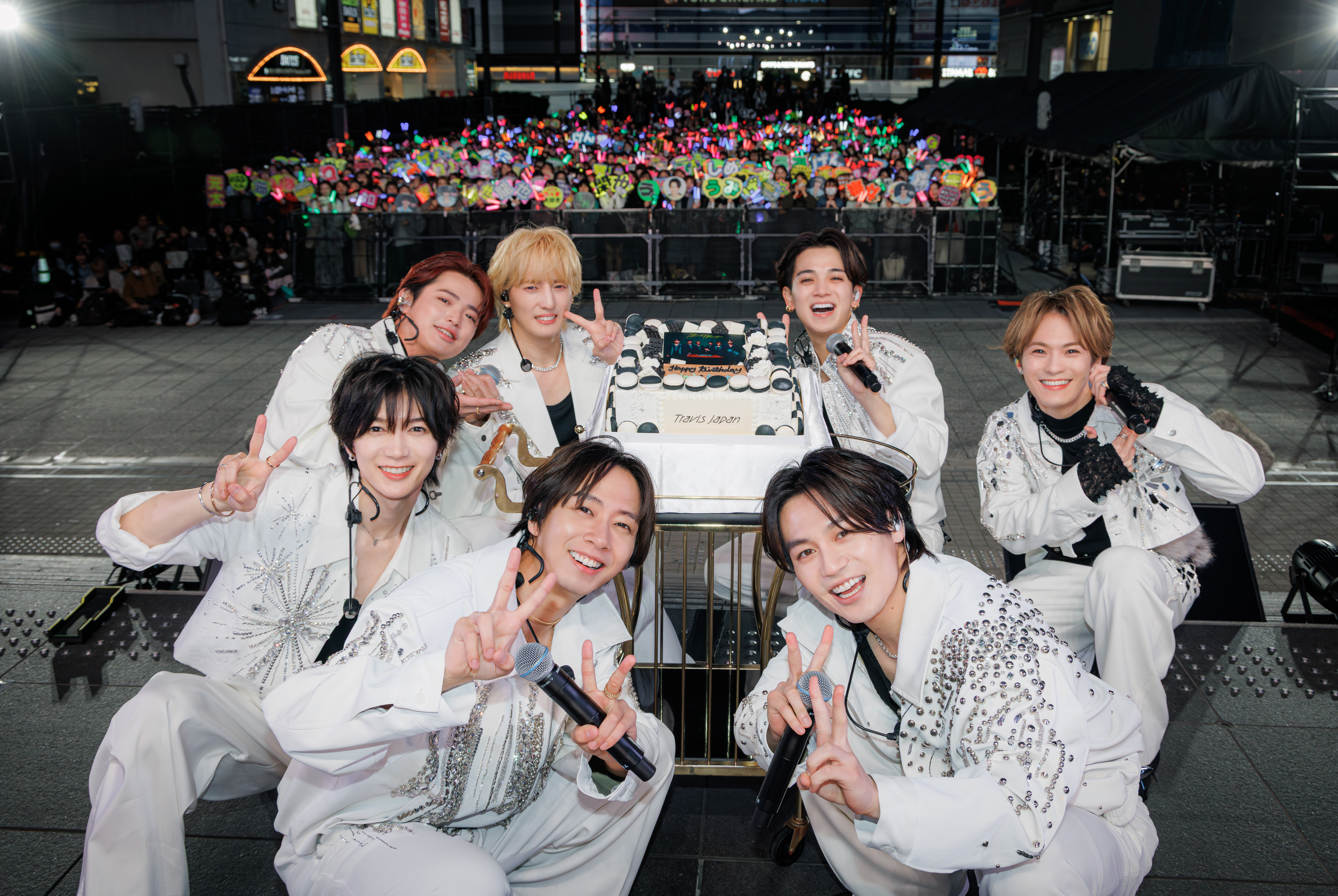 A group of seven performers wearing white outfits is celebrating with a cake that reads 'Travis Japan' in front of a cheering crowd holding colorful light sticks and signs.