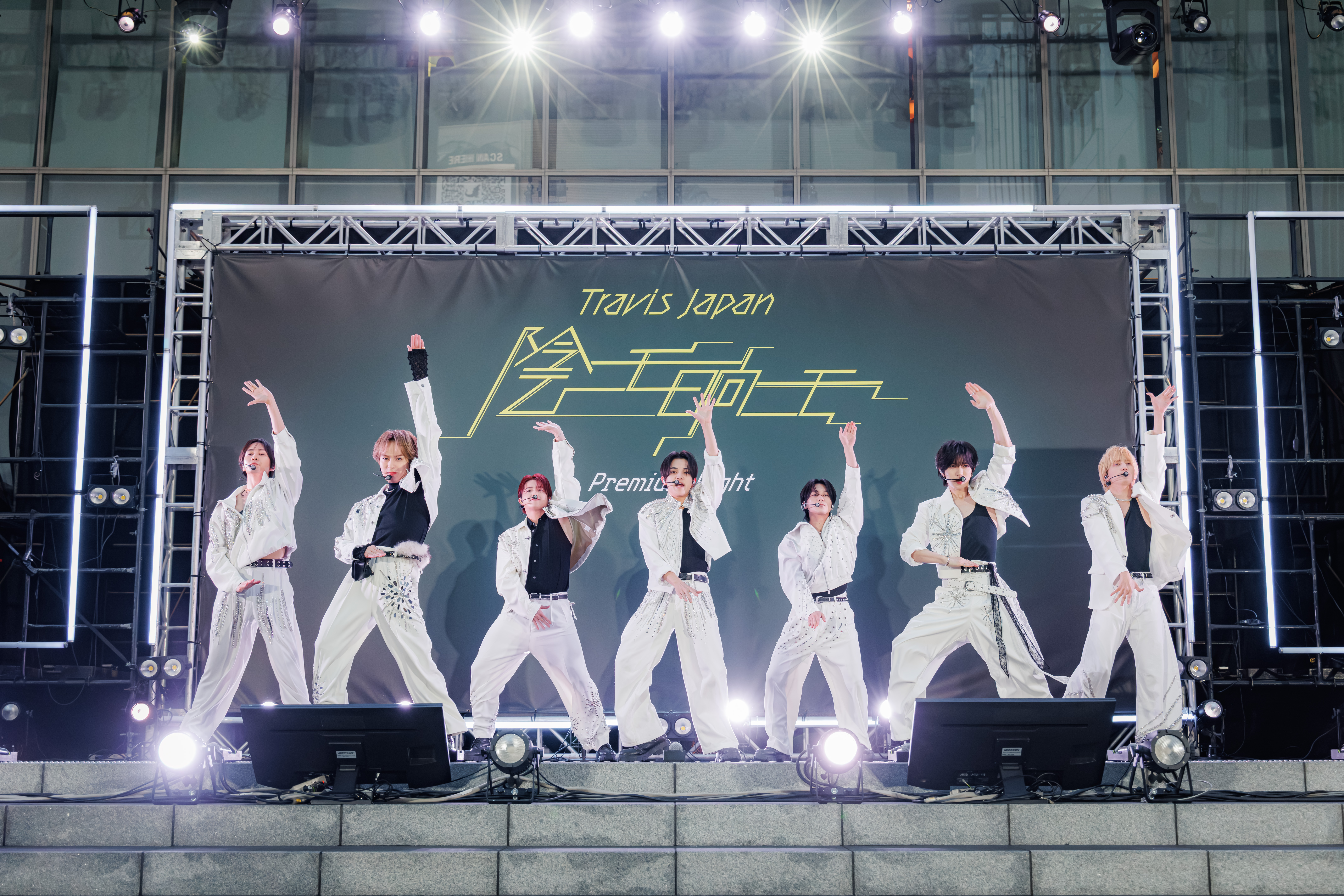 A performing boy band, Travis Japan, dressed in stylish outfits, showcases a dynamic dance routine on stage with a large backdrop displaying their name and event title.