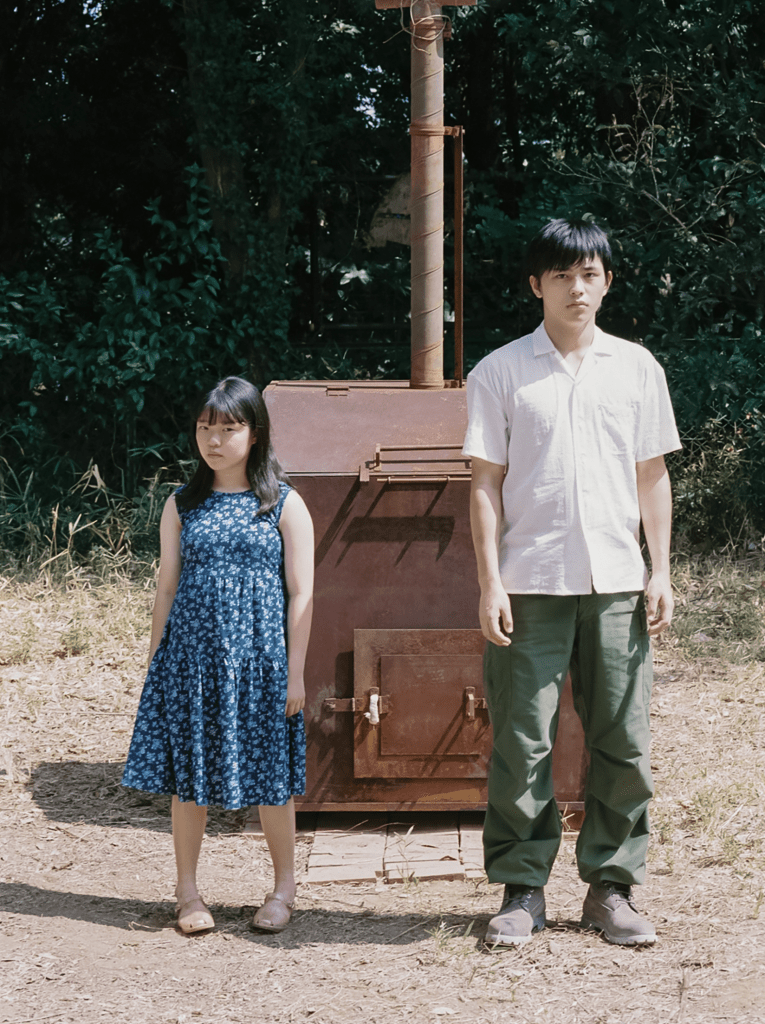 A young girl in a blue floral dress stands next to a teenage boy in casual clothing, both positioned near a metal structure in a wooded outdoor setting.