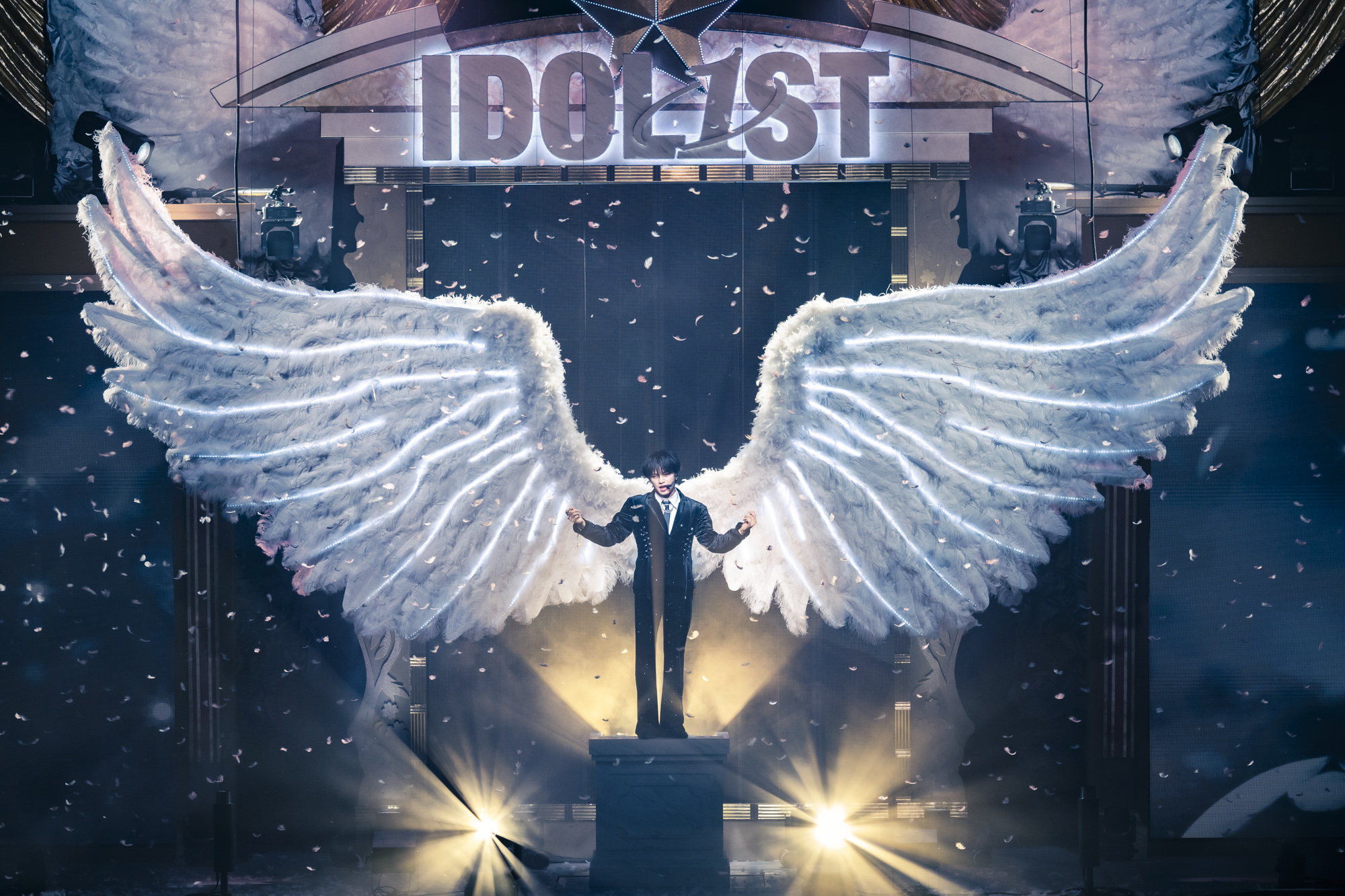 A performer stands on a pedestal with large, illuminated white angel wings, surrounded by swirling confetti and dramatic lighting, on a stage with the word 'IDOLIST' displayed above.