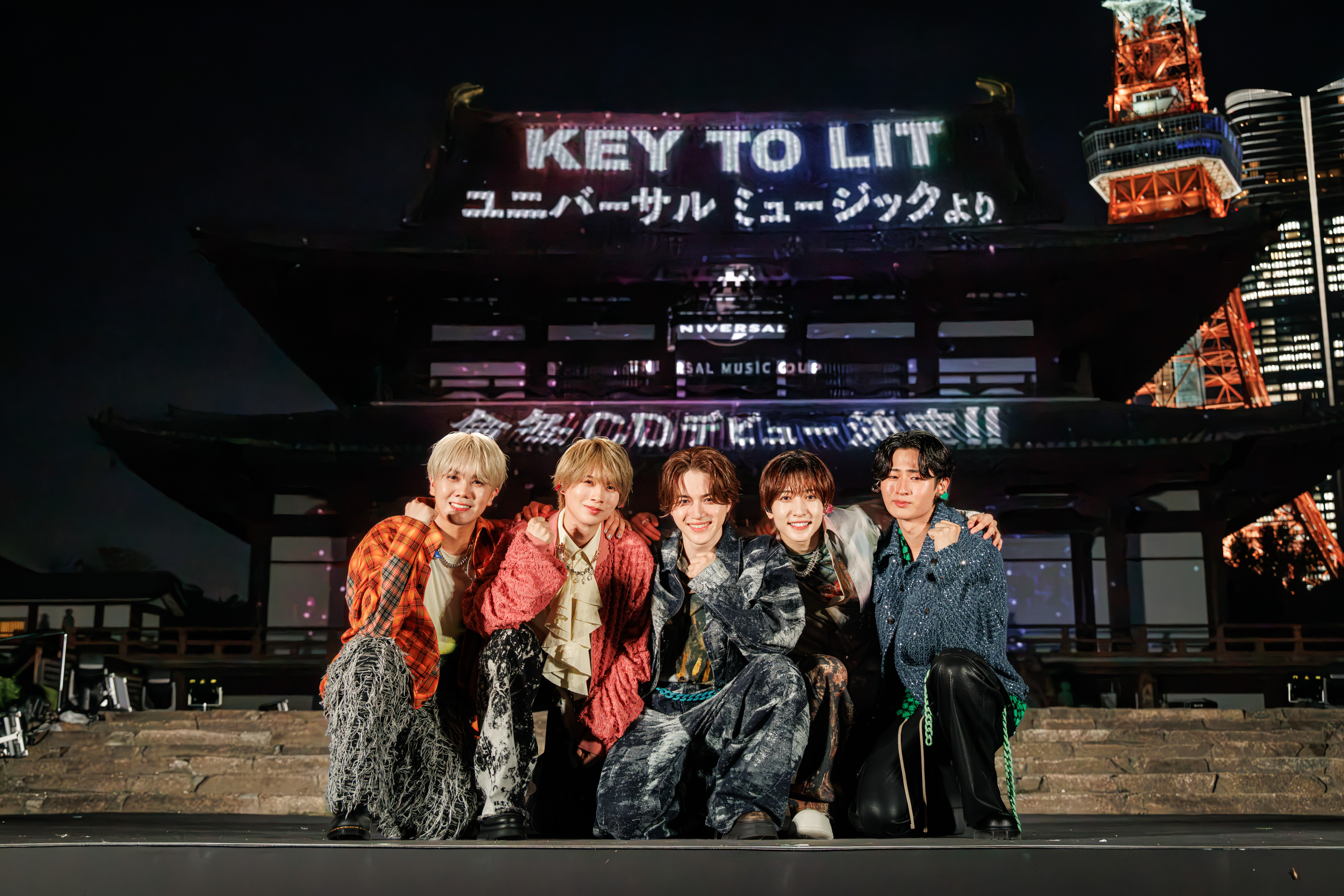 A group of five performers posing together in front of a pagoda-style structure illuminated with colorful lights, featuring the text 'KEY TO LIT' and 'UNIVERSAL MUSIC'. The scene takes place at night.