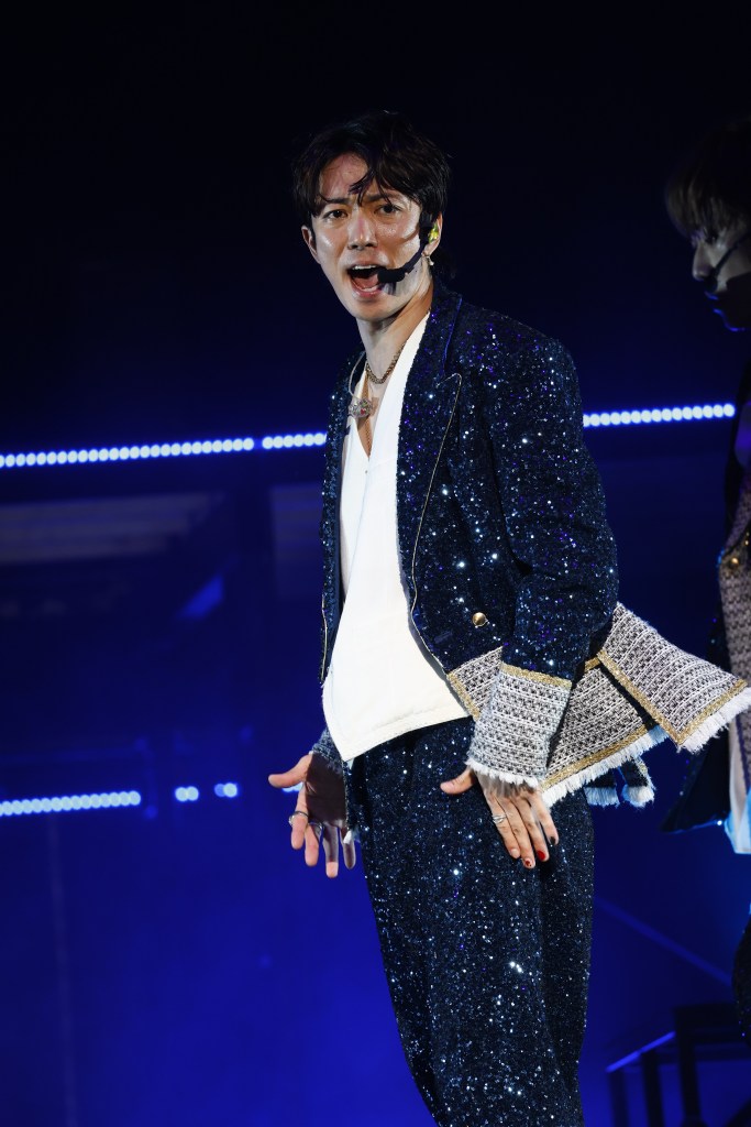 A performer wearing a sparkly blue suit and a white shirt, singing on stage with a microphone, illuminated by bright lights.