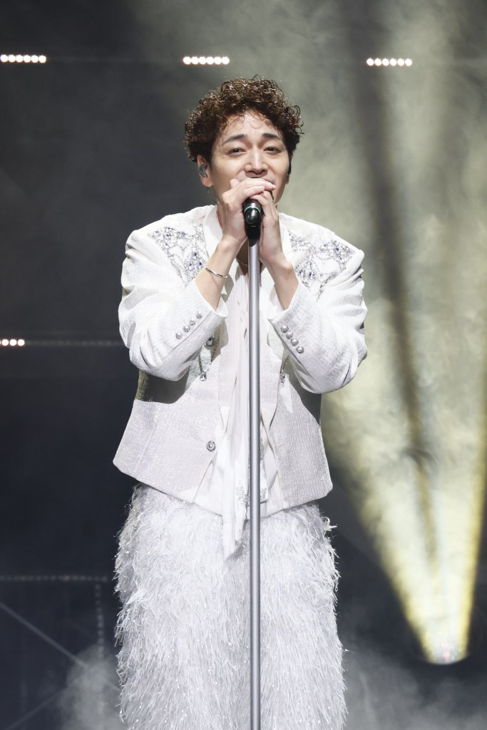 A male singer performing on stage, wearing a sparkling white suit and holding a microphone stand, illuminated by stage lights.