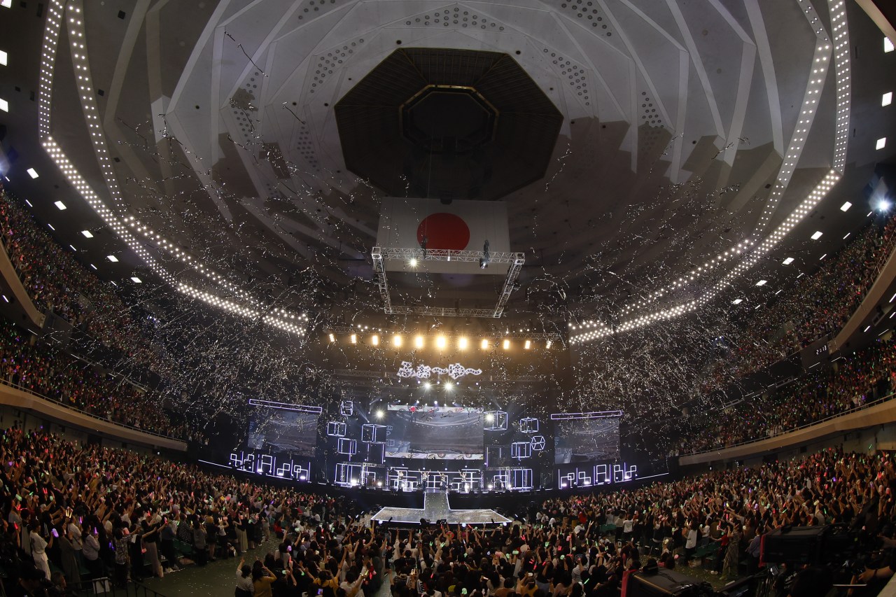 A large concert hall filled with excited fans celebrating during a live performance. Confetti is falling from the ceiling, and colorful lights are illuminating the crowd. A stage with bright lights is visible in the foreground, along with a large Japanese flag hanging above.