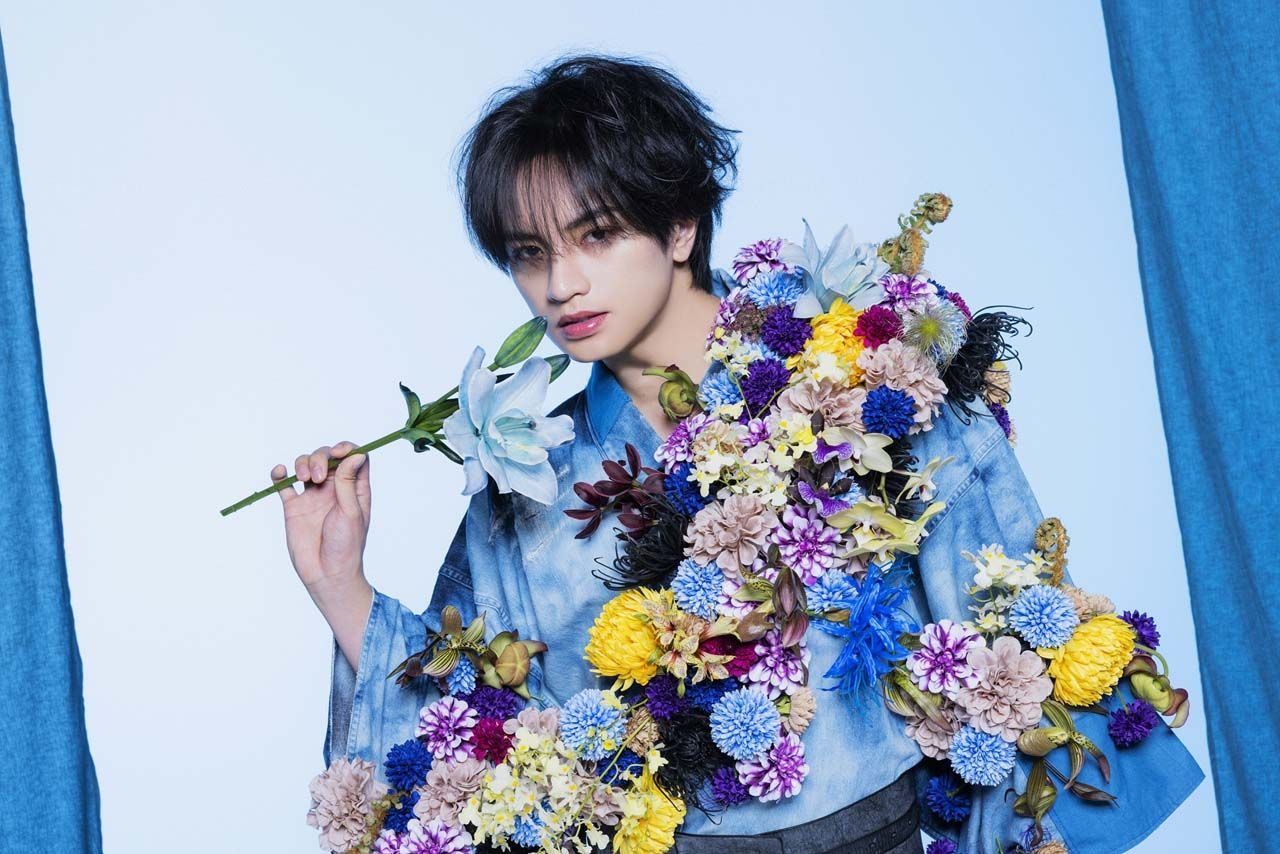 A person wearing a denim kimono adorned with colorful artificial flowers, holding a light blue flower in one hand and posing against a pastel blue backdrop.