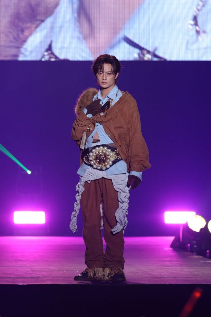 A model walking on stage in a unique outfit featuring a brown outer layer, a light blue shirt, and an embellished belt, with dramatic stage lighting in the background.
