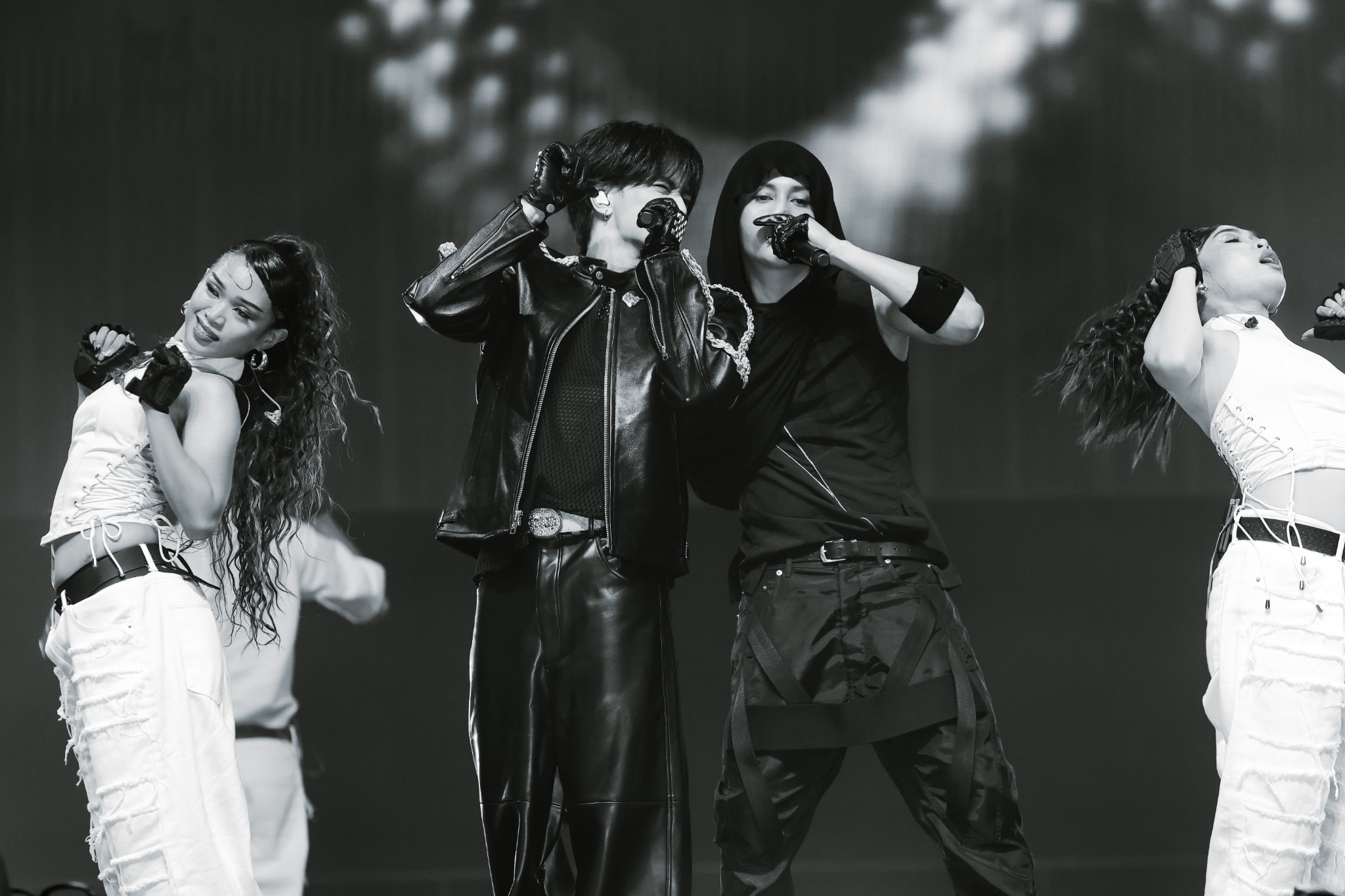 A monochrome image of a musical performance featuring two male singers and two female dancers, all wearing stylish outfits and performing energetically on stage.