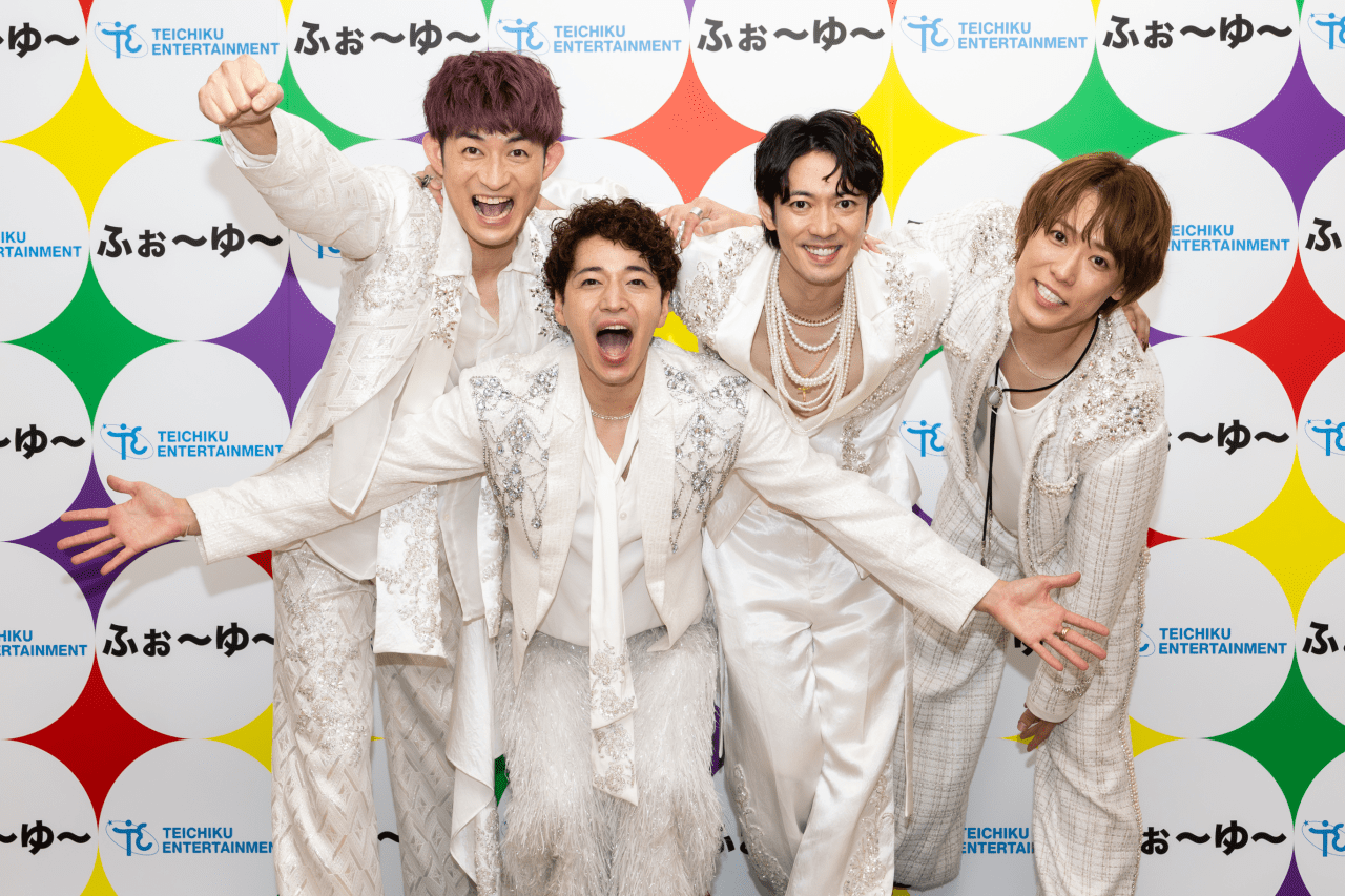 Four performers in white costumes joyfully posing together in front of a colorful backdrop with the Teichiku Entertainment logo.