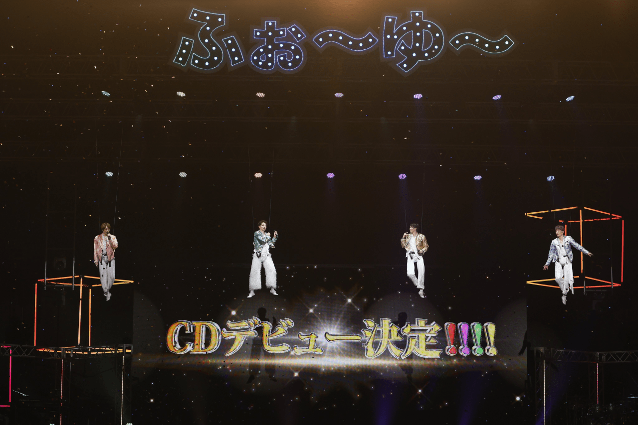 Four performers suspended in mid-air on stage, dressed in colorful outfits, with shiny decorations and a bright sign announcing their CD debut.