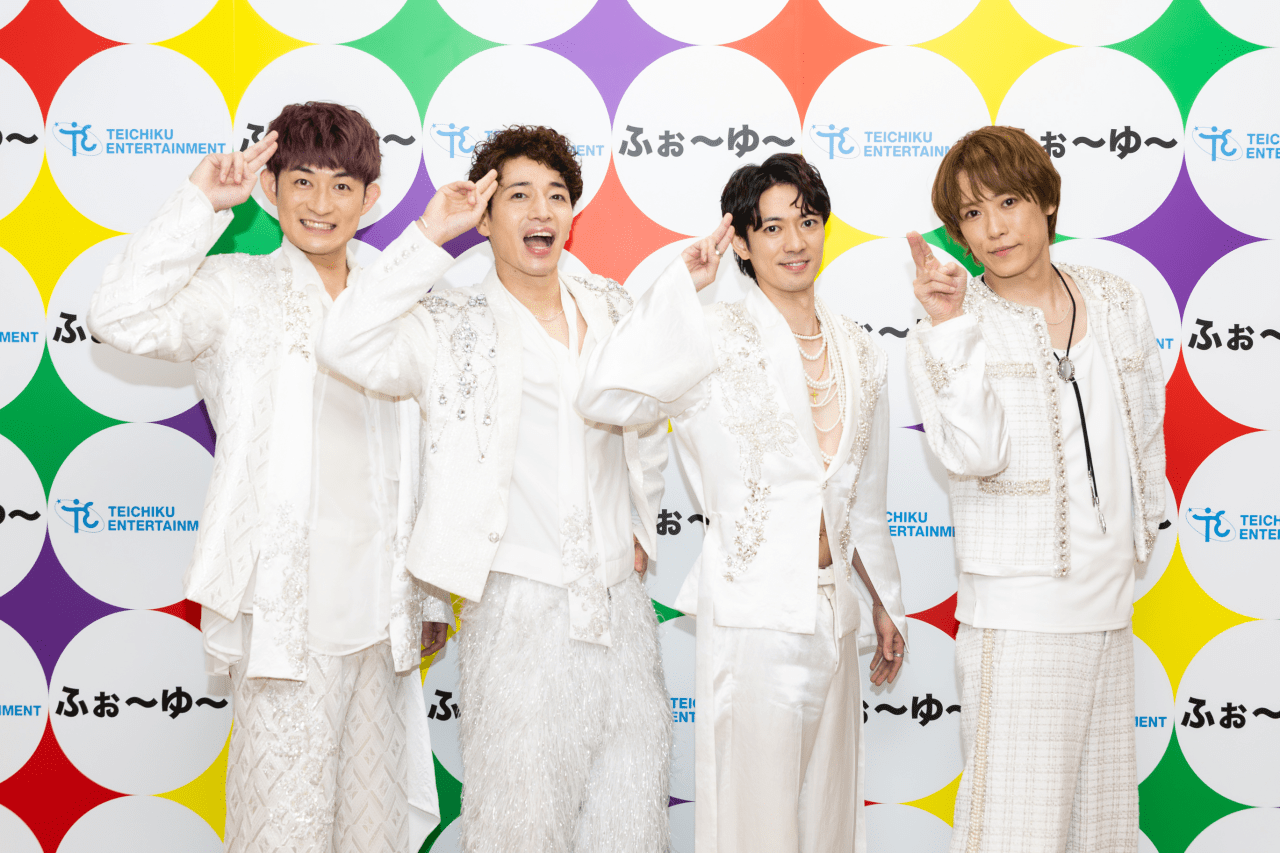 Four male performers in white, sparkly outfits posing playfully with their fingers raised in a saluting gesture against a colorful backdrop with circles and logos.