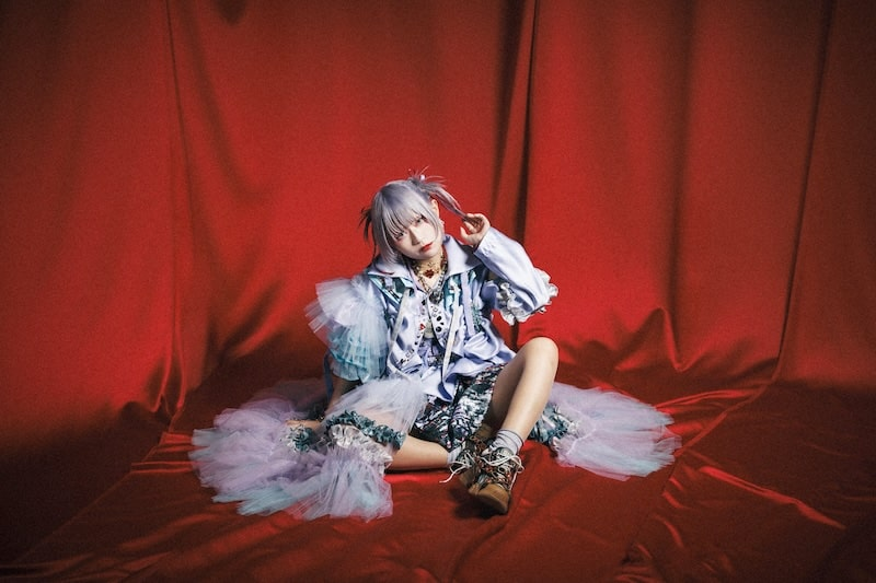 A young model in a colorful outfit with layers of tulle, sitting on a red draped surface, posing playfully with one hand near her face.