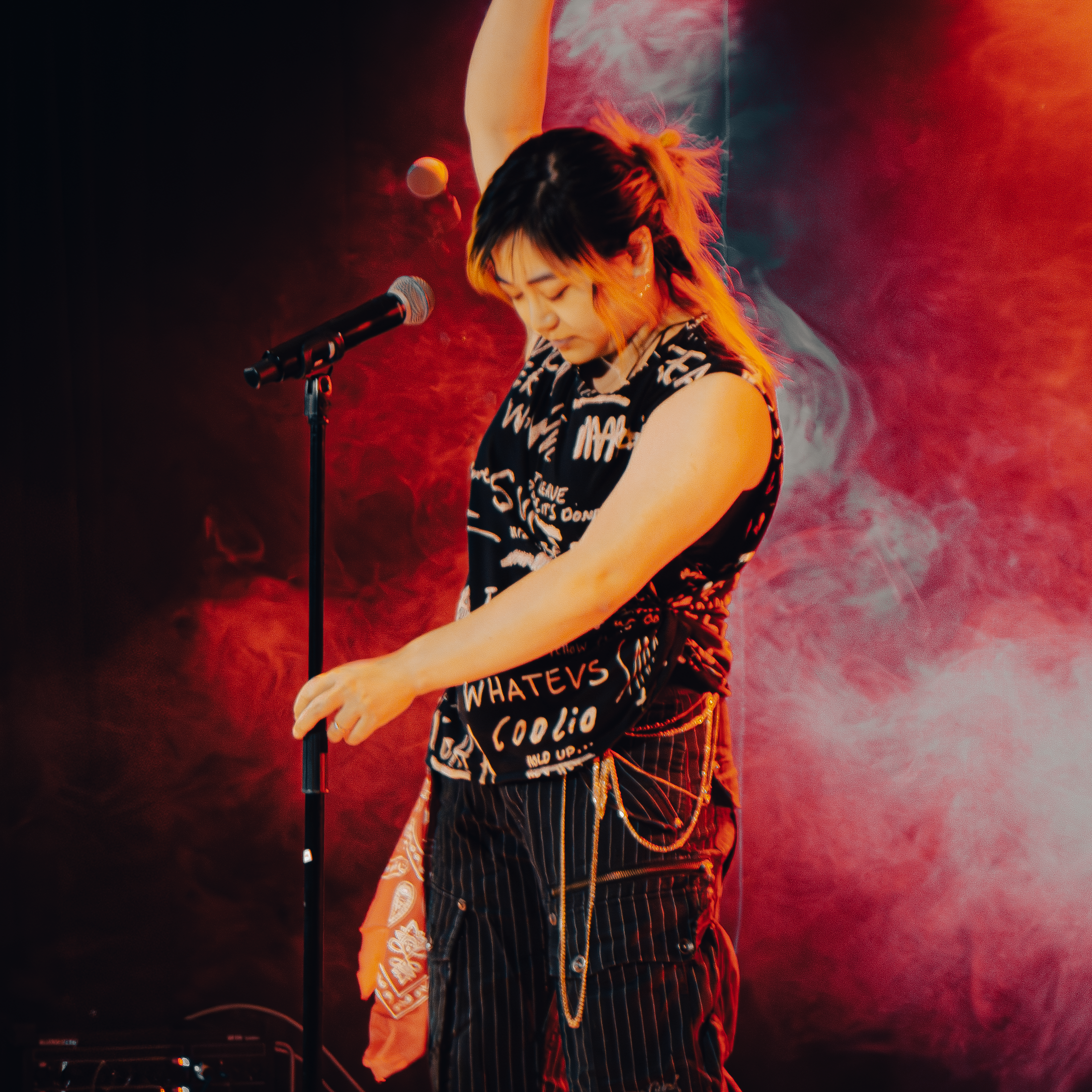A performer on stage, wearing a graphic tank top and striped pants, holds a microphone with one hand raised. The background features colorful lighting and smoke effects.