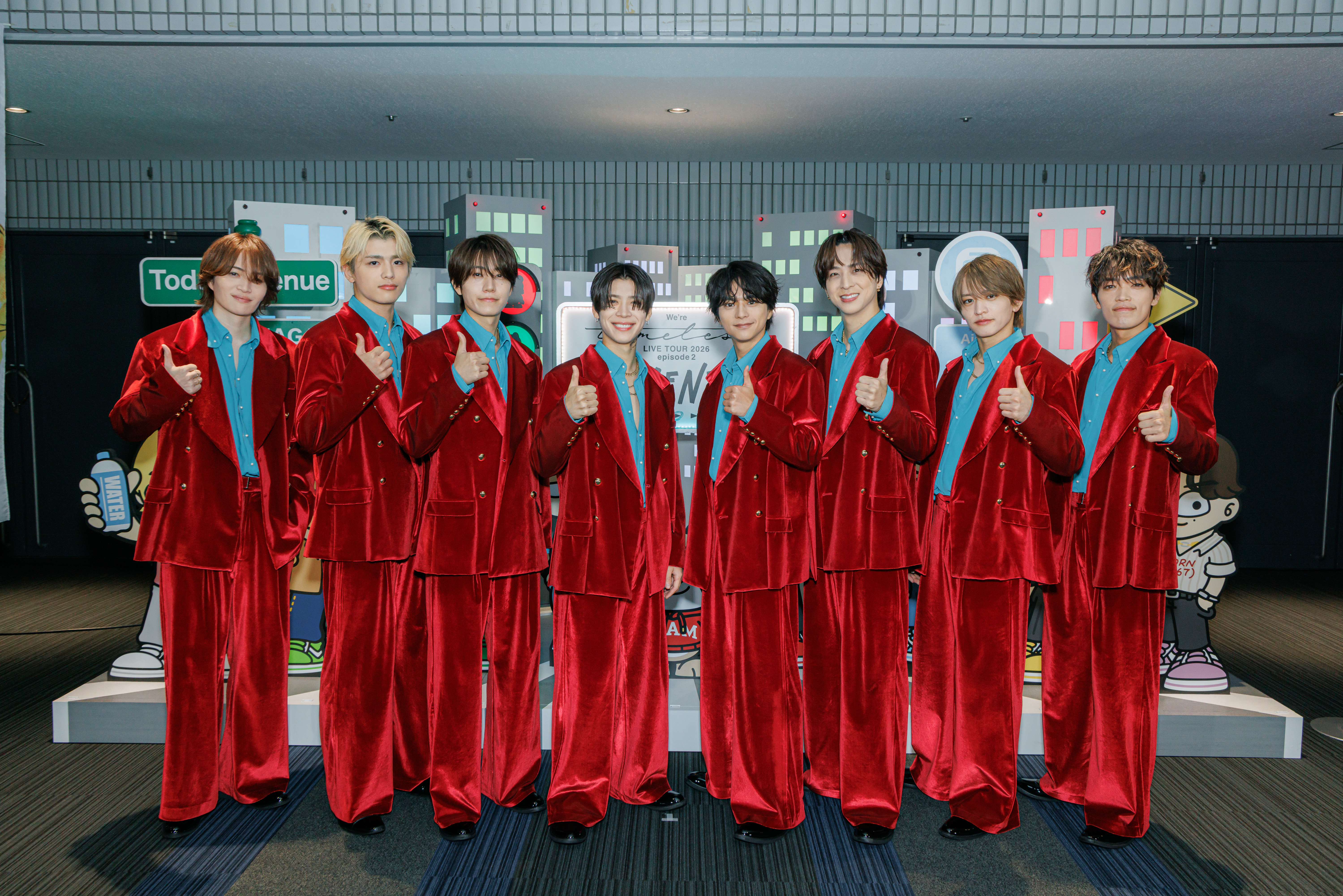 A group of nine individuals posing together, all wearing matching red velvet suits with blue shirts, giving thumbs up in a cheerful manner. The background features cartoon-like cityscapes and signs.